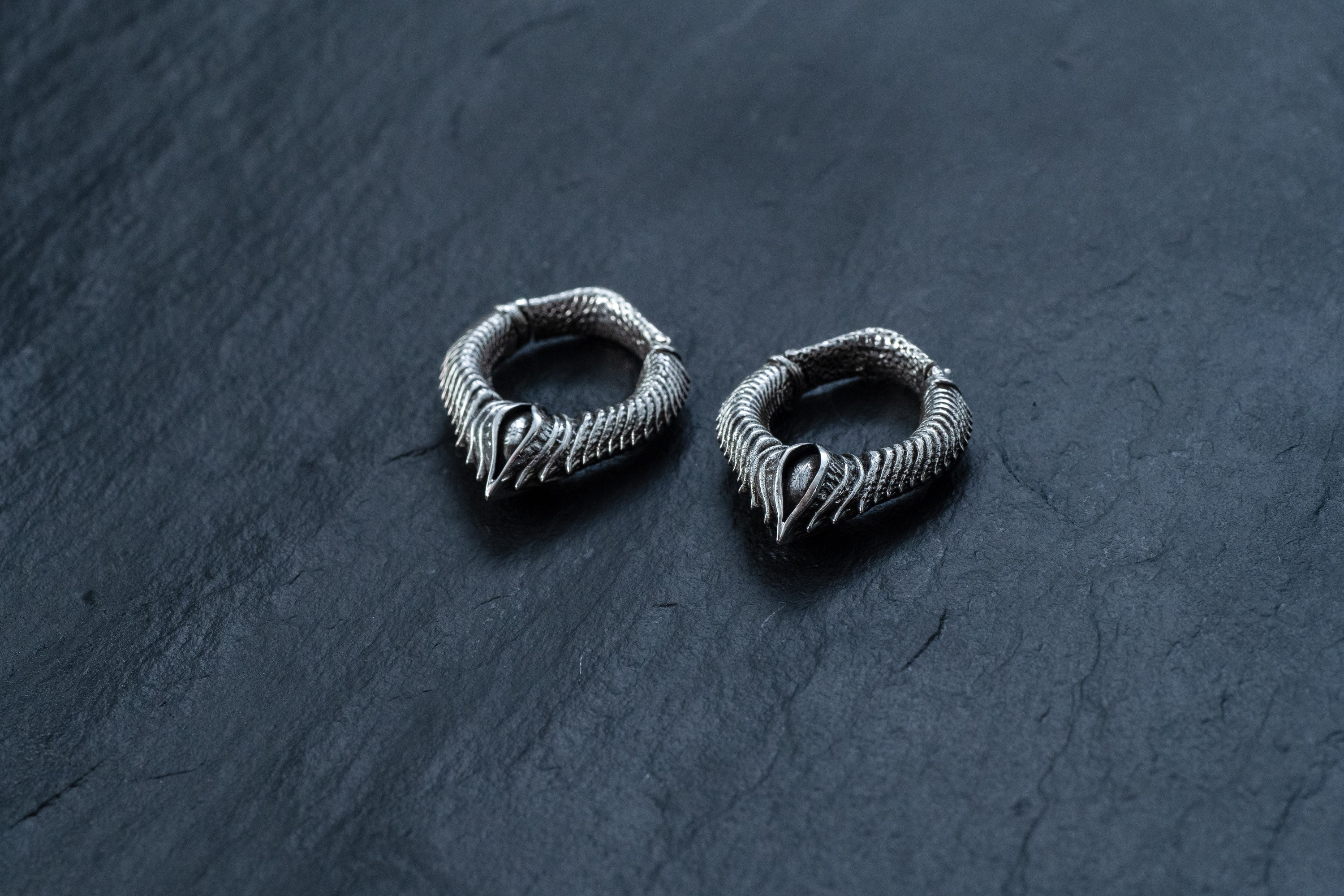 A pair of MARVDASHT Small Biomechanical Clicker Ear Weights in 925 silver, by Kaya Movement, are placed on a dark, textured surface. The 2-gauge ear weights feature an intricately detailed, scaly pattern with pointed ends, evoking a mythical, fantasy-inspired aesthetic with subtle sci-fi vibes.